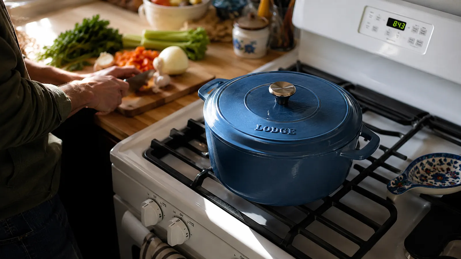 Lodge blue enamel Dutch oven sitting on a clean white stovetop in a bright, modest kitchen with a cutting board and fresh vegetables nearby